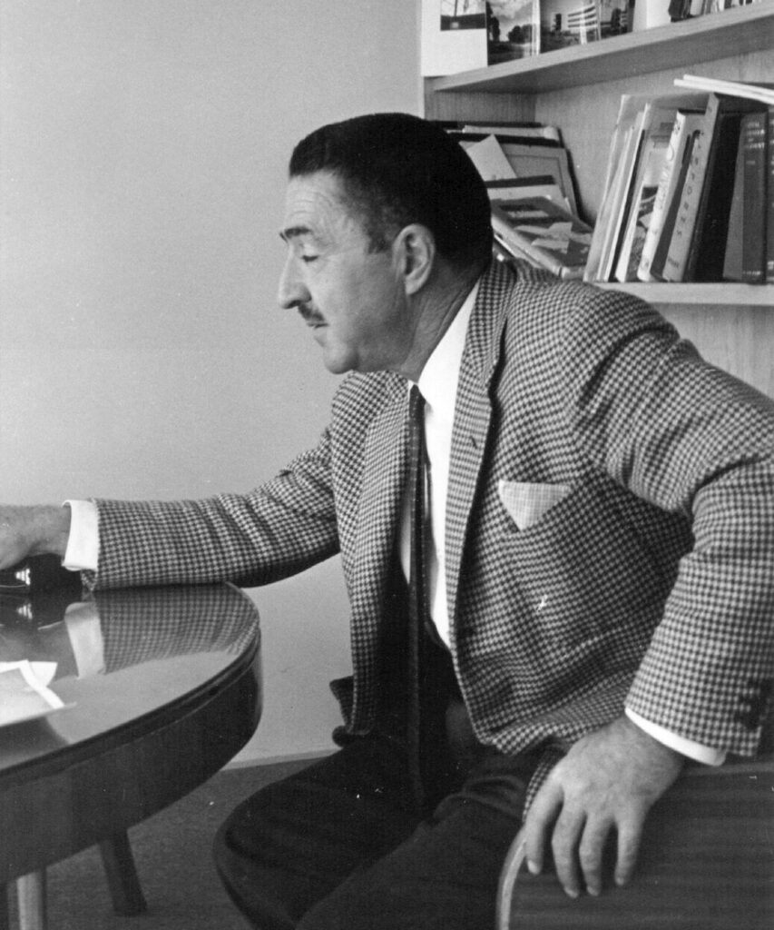 Retrato en blanco y negro del arquitecto español-mexicano Félix Candela sentado junto a una mesa de trabajo, con traje de cuadros y libros al fondo.