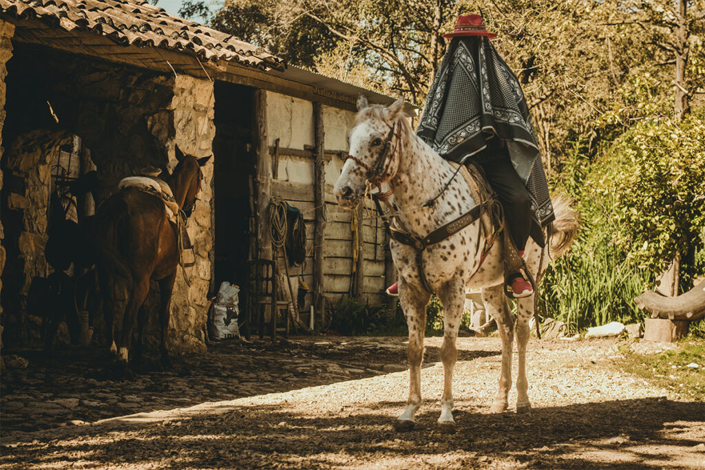 Doctor Nativo, cubierto con un poncho y sombrero rojo, monta un caballo en un entorno rural, evocando una figura enigmática que fusiona tradición campesina y estética contemporánea.