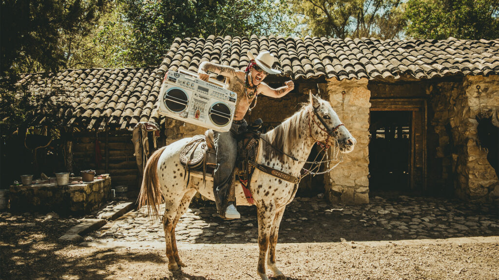 Doctor Nativo monta un caballo frente a una casa rural de piedra y teja, sosteniendo un radiocasete y levantando el puño con energía, en una escena que mezcla tradición, música y espíritu rebelde en un entorno campesino.
