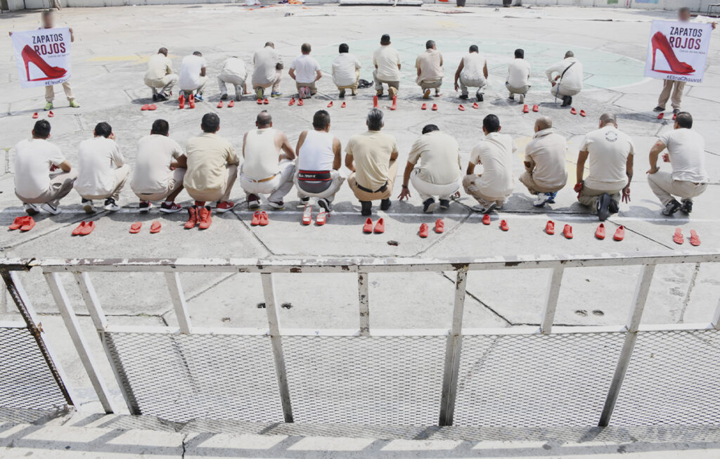 Image of the installation *Red Shoes*, in which a group of people crouching on the ground place pairs of red-painted shoes in an open space. This artistic action symbolizes the absence of women who have been victims of femicide and aims to raise social awareness about gender-based violence.