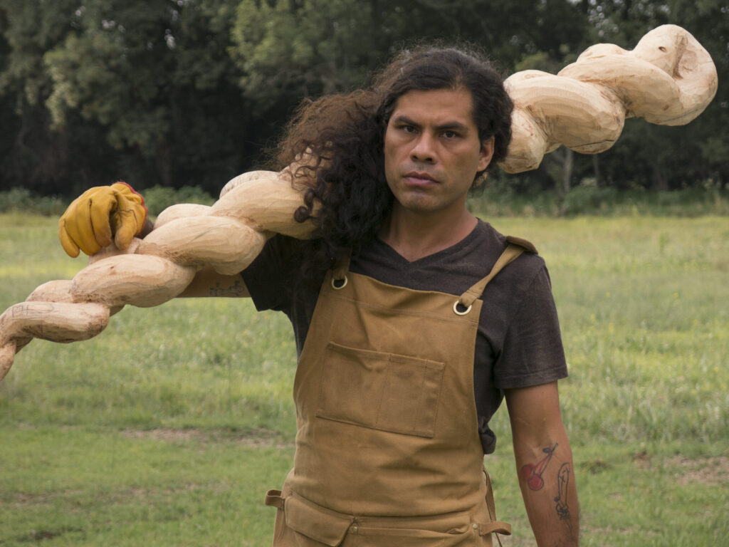 Artista de cabello largo y overol café sostiene una gran escultura de madera sobre el hombro en un campo al aire libre.