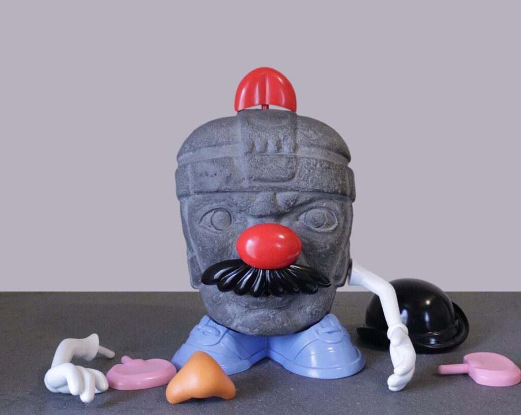 Escultura de cabeza olmeca intervenida con piezas del Señor Cara de Papa sobre fondo gris.