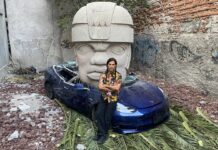 Chavis Mármol: «Aplasté un Tesla para trolear a Elon Musk» Artista sentado frente a un Tesla azul destruido por una enorme cabeza olmeca en un espacio urbano intervenido.