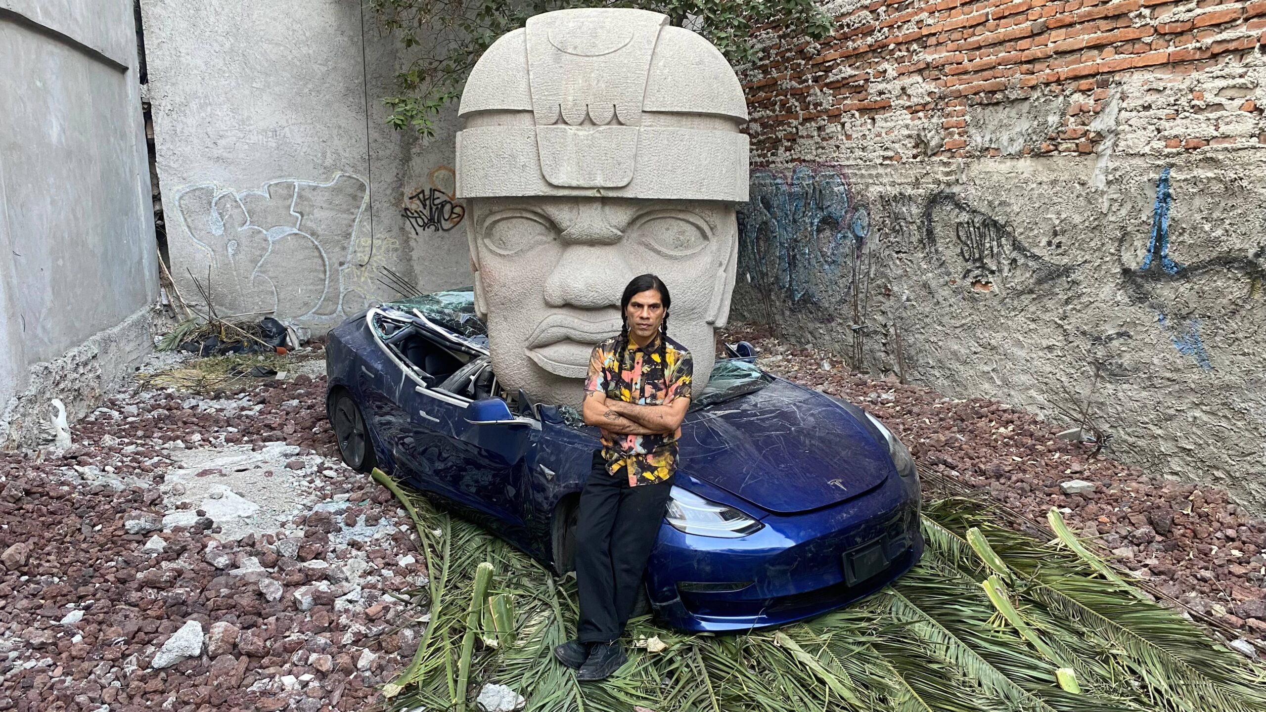 Artista sentado frente a un Tesla azul destruido por una enorme cabeza olmeca en un espacio urbano intervenido.