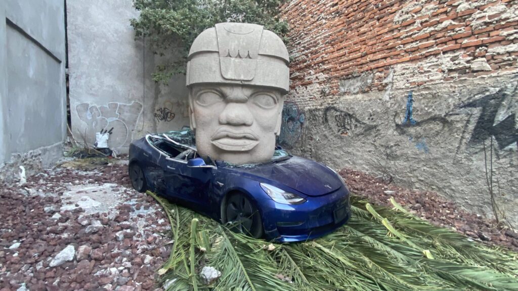 Gran cabeza olmeca aplasta un automóvil Tesla azul destruido en un terreno urbano rodeado de muros de ladrillo.