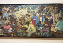 «México: ruta y destino» en el MUNAL: el arte que construyó una imagen idílica Mural colorido con vendedores, alimentos, flores y personajes en un mercado tradicional mexicano dentro de una sala de museo.