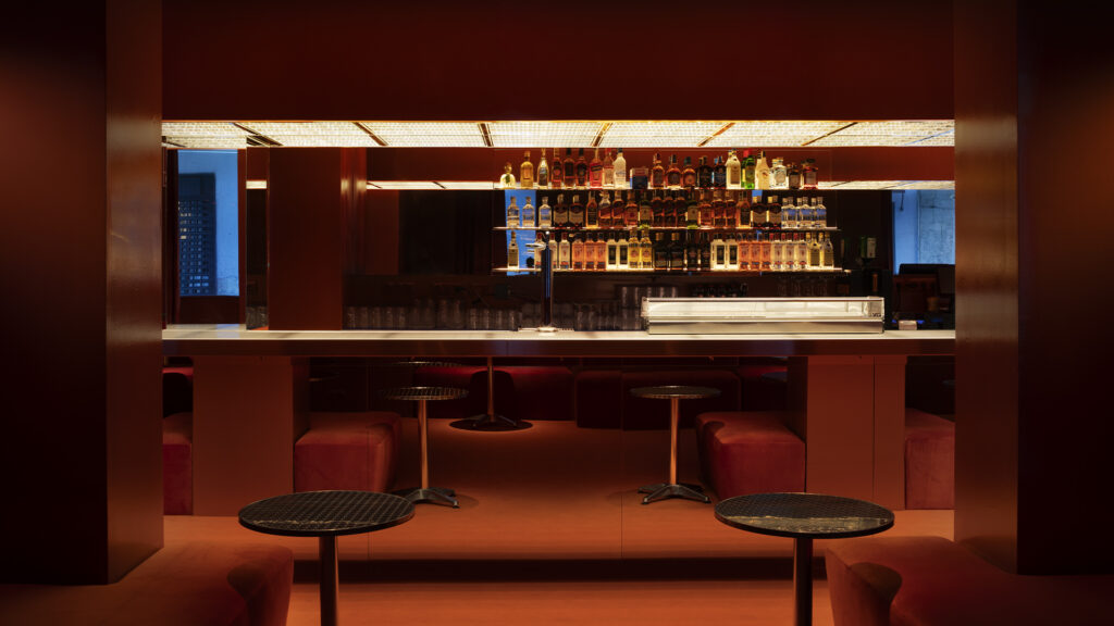 Bar de diseño contemporáneo completamente rojo, con iluminación cálida, mesas negras y botellas expuestas detrás de la barra.