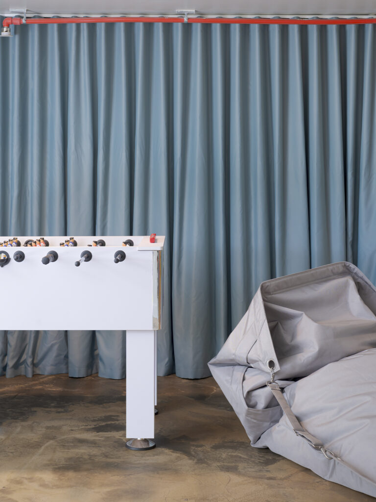 Espacio recreativo con futbolín blanco, cortinas azul grisáceo y puff de gran tamaño sobre suelo de hormigón.