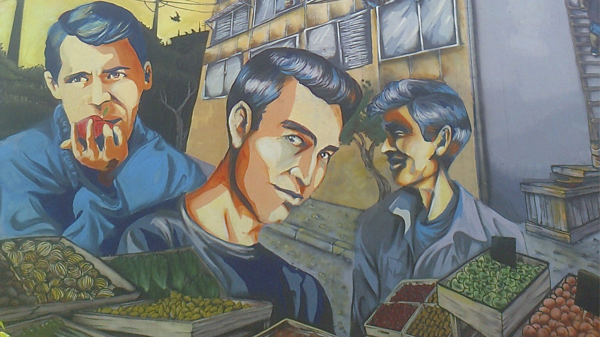 Mural dedicado a Los Prisioneros en un edificio de barrio, con retratos estilizados de los integrantes de la banda chilena frente a cajas de frutas y verduras, bajo un cielo anaranjado al atardecer.