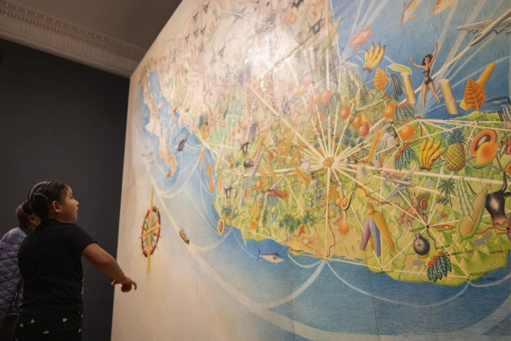 Niña observando un enorme mural cartográfico colorido de México en una sala oscura de museo.