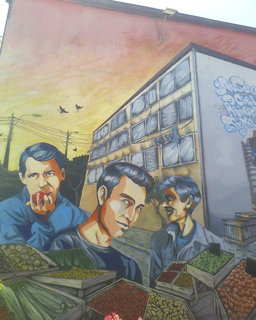 Mural dedicado a Los Prisioneros en un edificio de barrio, con retratos estilizados de los integrantes de la banda chilena frente a cajas de frutas y verduras, bajo un cielo anaranjado al atardecer.