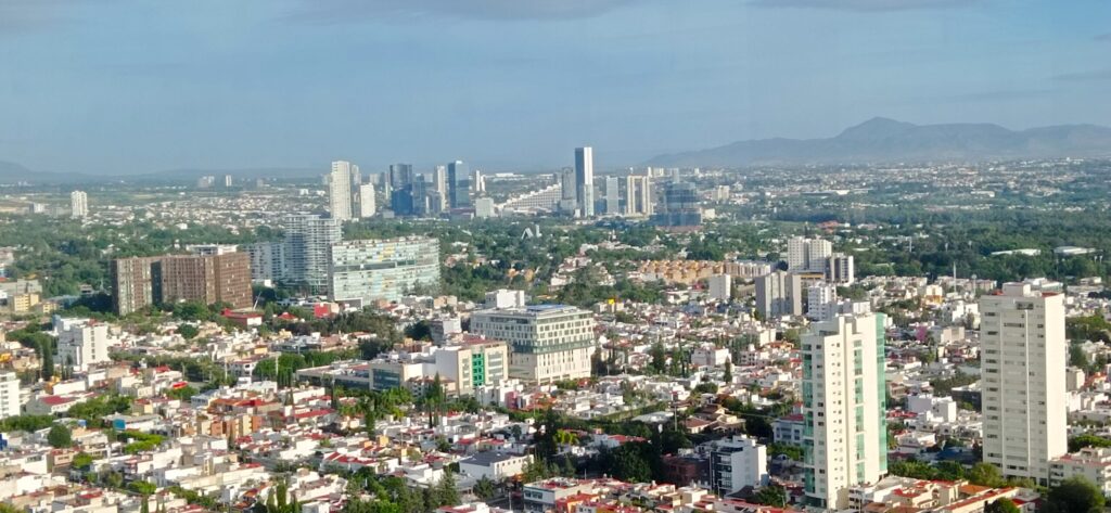 Una vista panorámica de la ciudad de Guadalajara, México, desde una perspectiva de dron hacia el noroeste. La ciudad se extiende hasta el horizonte con rascacielos y edificios comerciales bajo un cielo despejado.