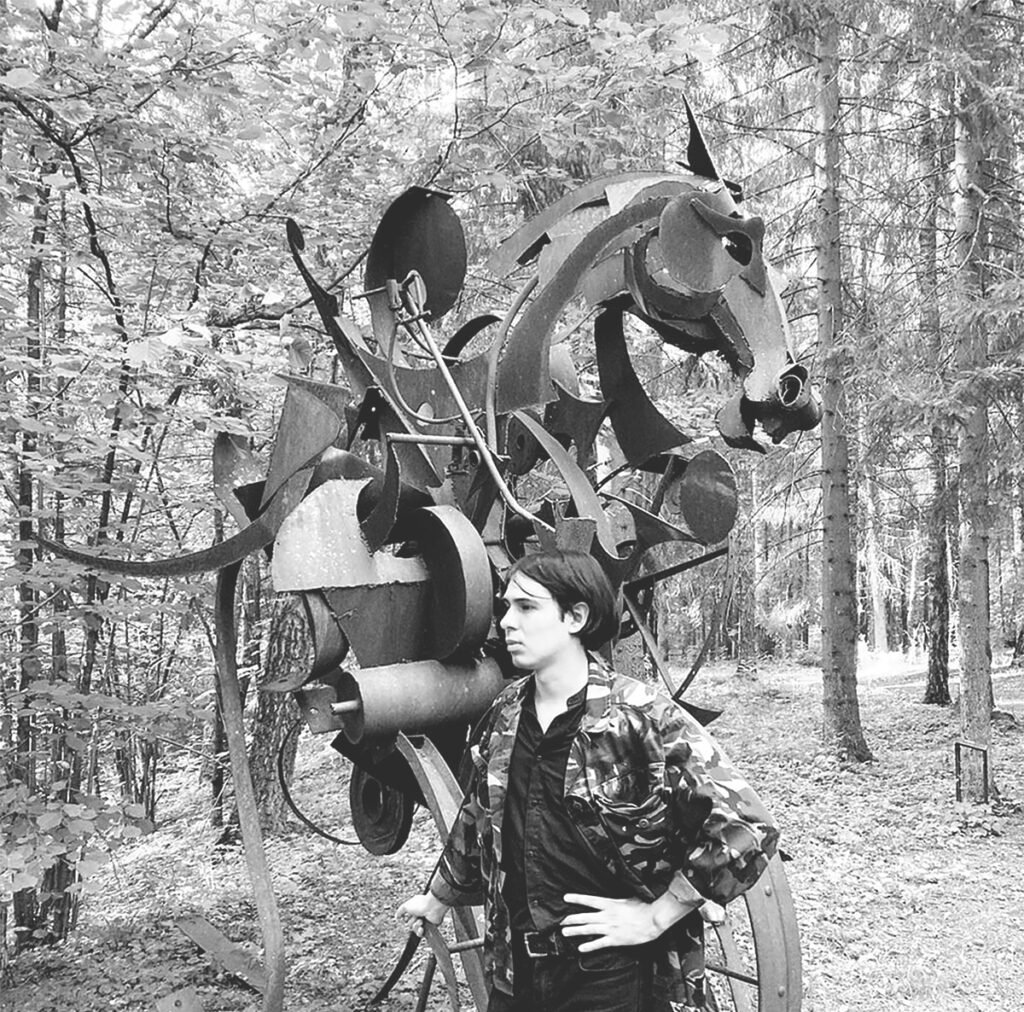 Rogelio Serrano posa junto a una escultura metálica abstracta en un bosque europeo.