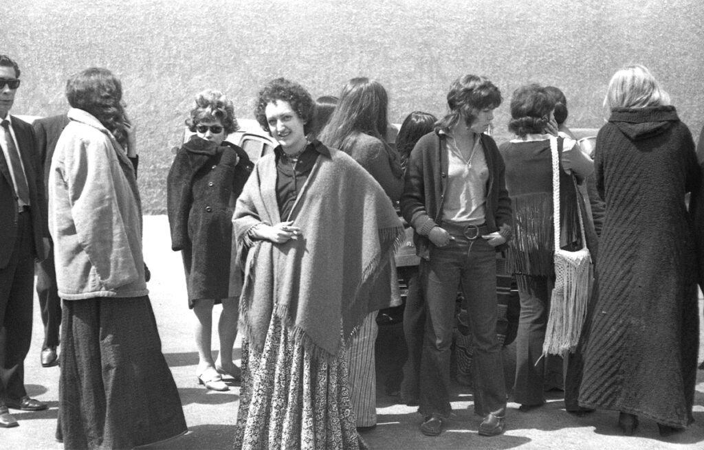 Fotografía en blanco y negro de varias mujeres jóvenes detenidas tras la redada de 1971, esperando de pie en un patio exterior. Visten ropa de la época contracultural, incluyendo ponchos, faldas largas estampadas y pantalones de campana.