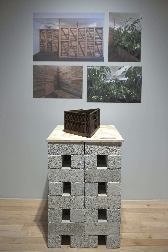 Vista de una sección de la exposición con una maqueta de madera sobre un pedestal hecho de bloques de concreto gris. Al fondo, en la pared gris, se observan cuatro fotografías que muestran estructuras de madera y vegetación integradas a la arquitectura.