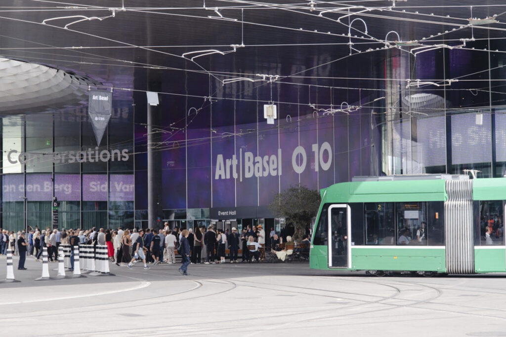 Fachada vidriada del pabellón de Art Basel con un letrero digital de la sección Zero 10 y un tranvía verde circulando por delante en las calles de Basilea.