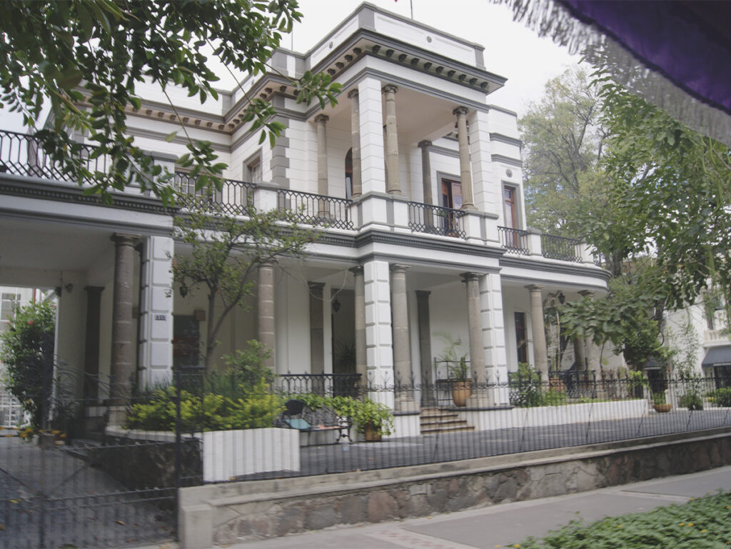 Una toma a nivel de calle de una gran casa de dos pisos con una fachada de estuco blanco y columnas de piedra estriadas que sostienen balcones con barandillas de hierro forjado.
