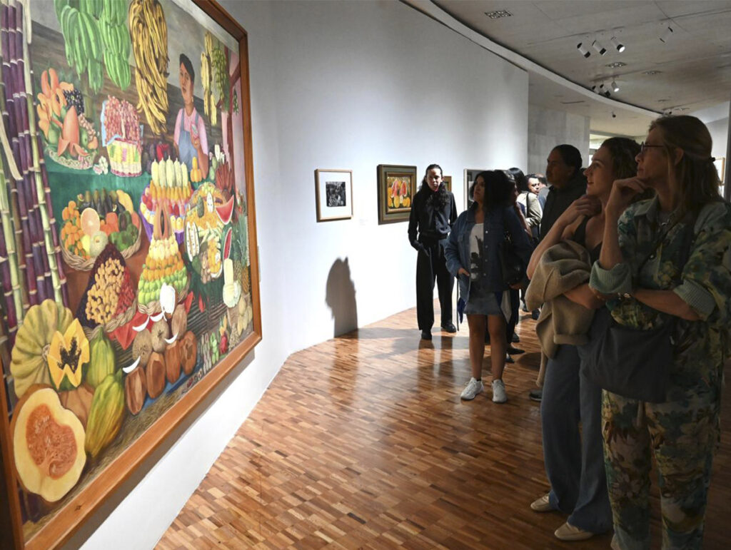 An angled view of a room in the Museum of Modern Art with a wooden floor. In the foreground, on the left, a large-scale painting stands out, filled with colorful tropical fruits, sugarcane, and a vendor.