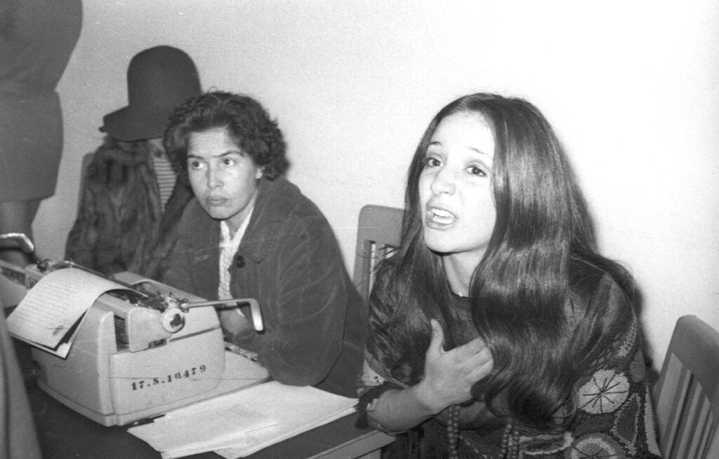 Fotografía en blanco y negro en un interior de oficina policiaca. Isela Vega, con el cabello largo y suelto y un huipil o blusa artesanal bordada, aparece hablando intensamente con la mano en el pecho frente a una máquina de escribir, mientras otra mujer observa al fondo.