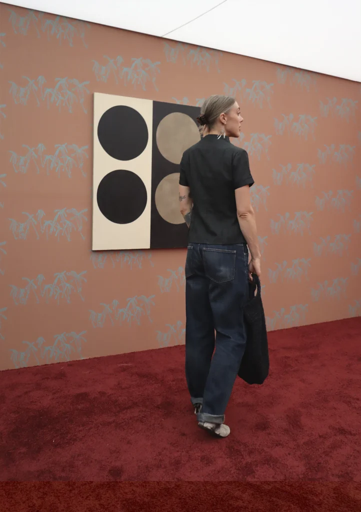 Una mujer de espaldas con cabello recogido y jeans contempla un cuadro cuadrado dividido en dos bloques —blanco y negro— con círculos concéntricos, montado sobre una pared rosa pastel con dibujos de palmeras.