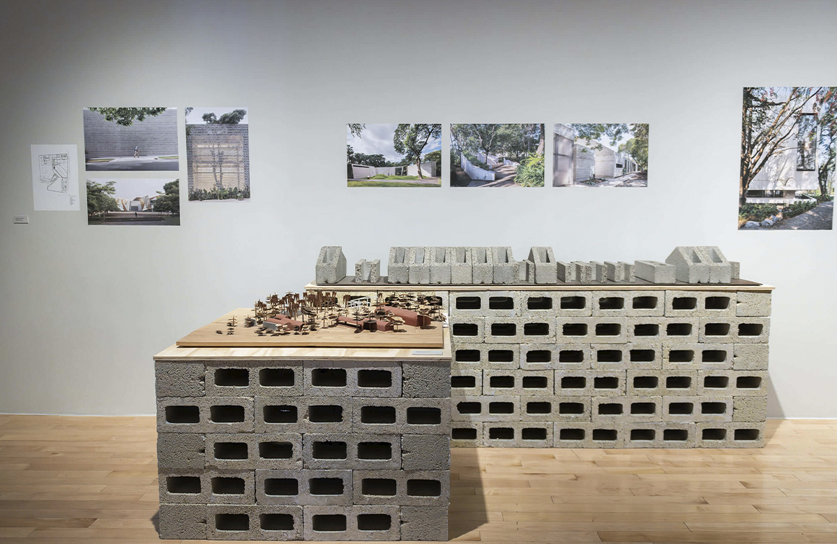 Amplia sección de la galería que exhibe maquetas sobre mesas construidas con bloques de construcción perforados. La pared blanca del fondo contiene una serie de planos arquitectónicos y fotografías de proyectos construidos en México durante las últimas décadas.