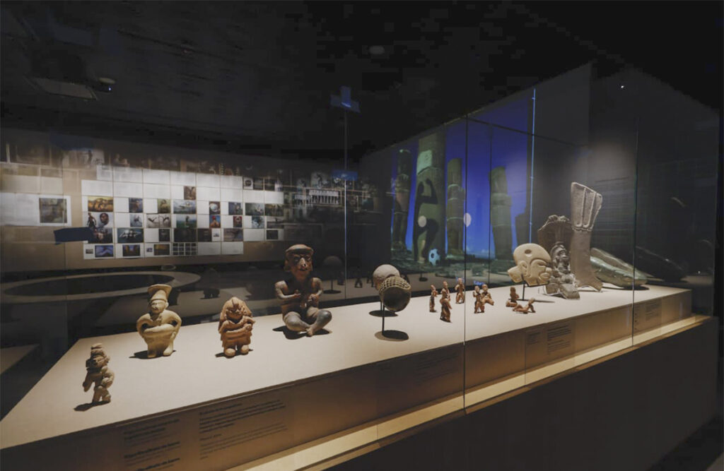Vista de una sala oscura del Museo Nacional de Antropología donde se exhiben piezas arqueológicas vinculadas al juego de pelota mesoamericano frente a fotografías y proyecciones de la exposición Futbol 2026. Annie Leibovitz.