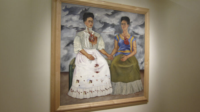 Fotografía en primer plano y de frente de la célebre pintura modernista 'Las dos Fridas' de Frida Kahlo, enmarcada en madera clara y colgada sobre un muro beige texturizado de la galería.