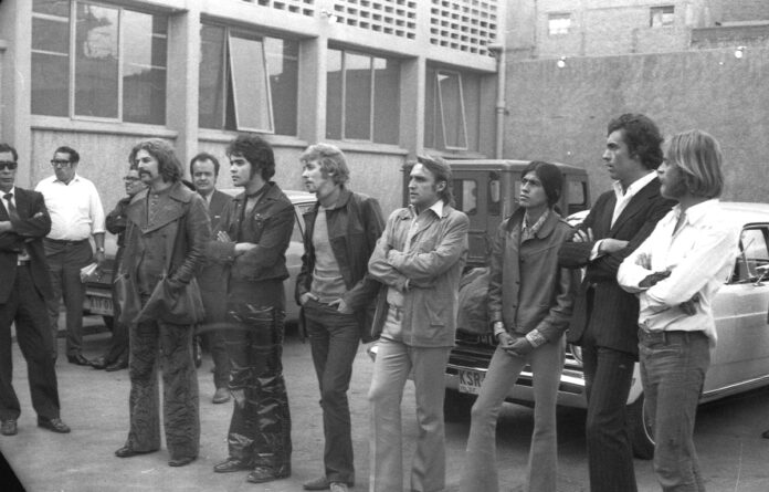 Esta fotografía en blanco y negro, con grano y alta contraste, captura una escena caótica y llena de tensión en un estacionamiento exterior de asfalto en Ciudad de México, durante la década de 1970.