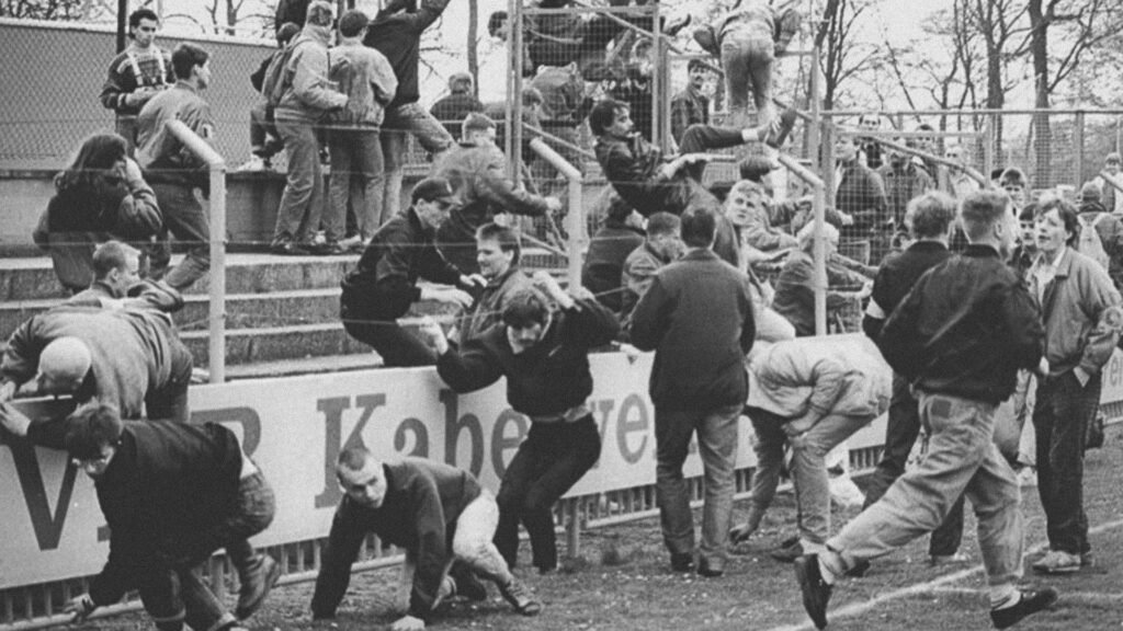 Fotografía en blanco y negro de decenas de aficionados saltando las bardas e invadiendo un campo de futbol durante un disturbio; varias personas corren, trepan las rejas y se aglomeran junto a las gradas.