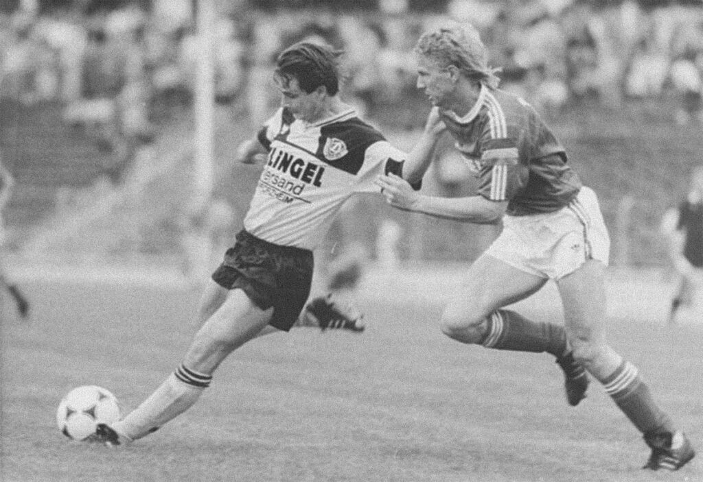 Fotografía en blanco y negro de Jorge Valdano disputando un balón con un defensor durante un partido de futbol, mientras ambos corren sobre el terreno de juego.