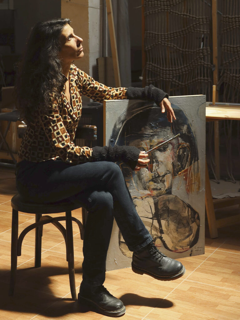 Eulogia Merle, artista argentina afincada en Madrid, sentada en su estudio junto a uno de sus retratos de gran formato mientras sostiene un pincel y observa hacia arriba.