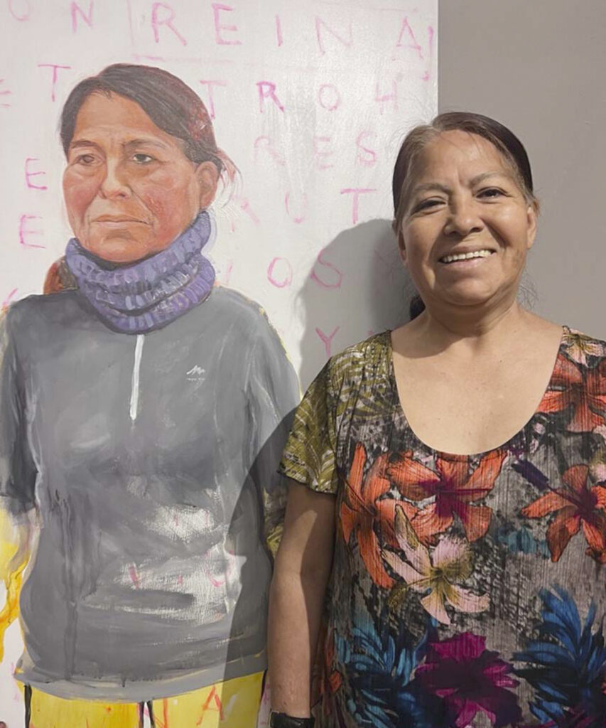 Dionisia junto al retrato que forma parte de Cama Adentro, la serie de Eulogia Merle que da rostro a trabajadoras domésticas latinoamericanas en España y reivindica sus historias de vida, migración y cuidados.