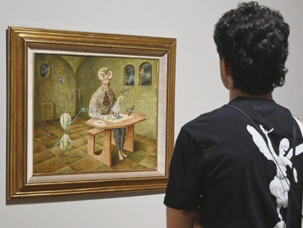A young man, seen from behind, is closely examining a surrealist painting with a gold frame by the artist Remedios Varo. The work depicts a figure with an owl's face in an alchemical workshop, surrounded by mystical artifacts and birds.
