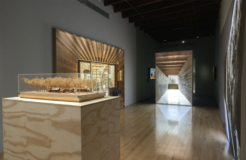 Perspectiva general de una sala de exhibición con pisos de madera clara. En primer plano destaca una maqueta en su pedestal y al fondo se observa un gran panel fotográfico que simula un túnel o pasillo de concreto que dirige la mirada hacia el fondo de la galería.
