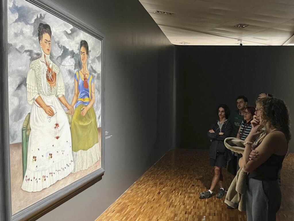 A perspective view of an exhibition hall with dark gray walls. In the background, a group of four people is intently viewing Frida Kahlo’s iconic painting “The Two Fridas,” which is illuminated by a spotlight.