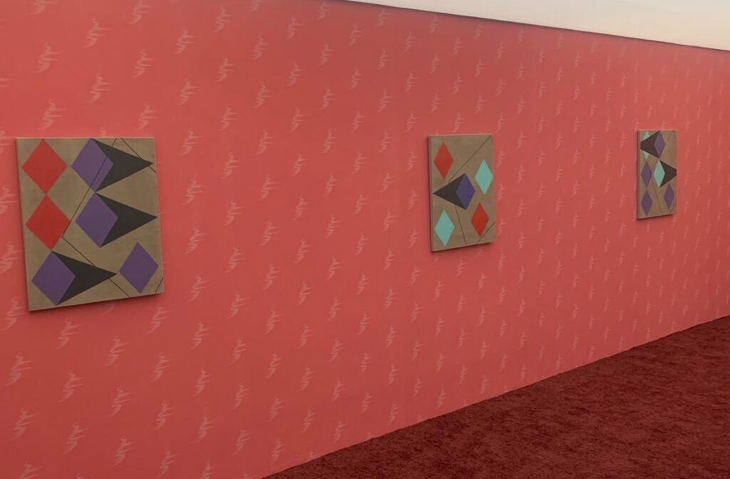 Vista de tres cuadros medianos montados en una pared verde oliva con sutiles patrones repetitivos. Los lienzos muestran diseños geométricos abstractos con picos en tonos naranja, amarillo flúor y marrón.