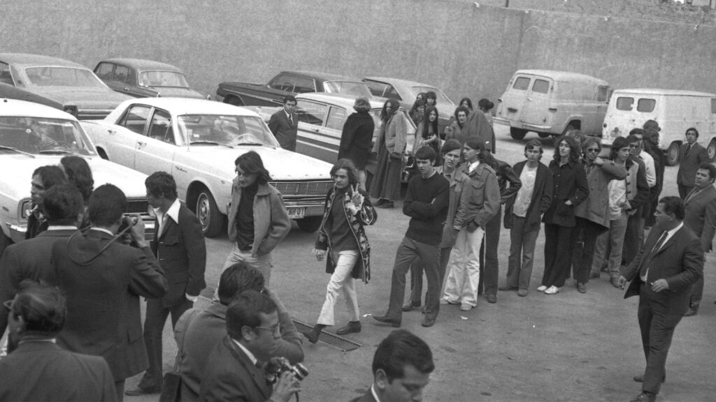 Fila de hombres jóvenes detenidos de pie en un estacionamiento exterior junto a vehículos de los años setenta.