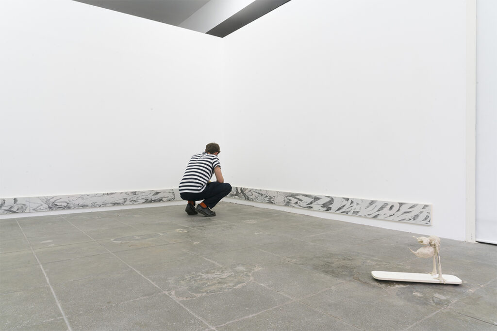 Persona agachada observando una instalación artística en una sala de exposición blanca; en primer plano se aprecia una pequeña escultura sobre una base baja.