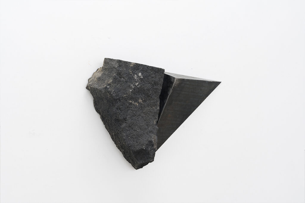 Escultura mural formada por una roca oscura integrada a una estructura geométrica de metal, instalada sobre una pared blanca en una galería de arte contemporáneo.