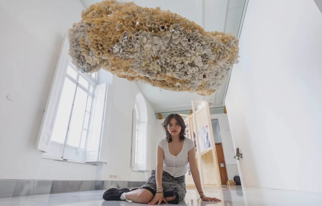 Nebraska Flores Espín sentada bajo una gran instalación escultórica suspendida de papel durante una exposición de arte contemporáneo.
