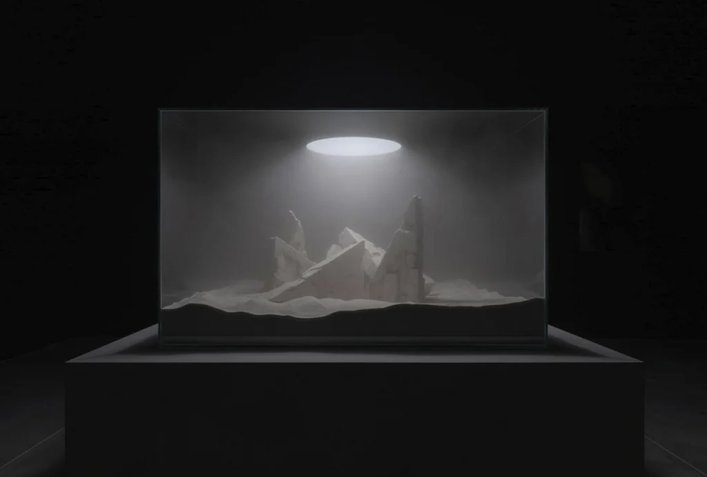 Vitrina iluminada desde el techo con estructuras geométricas semienterradas en arena blanca sobre un pedestal negro.
