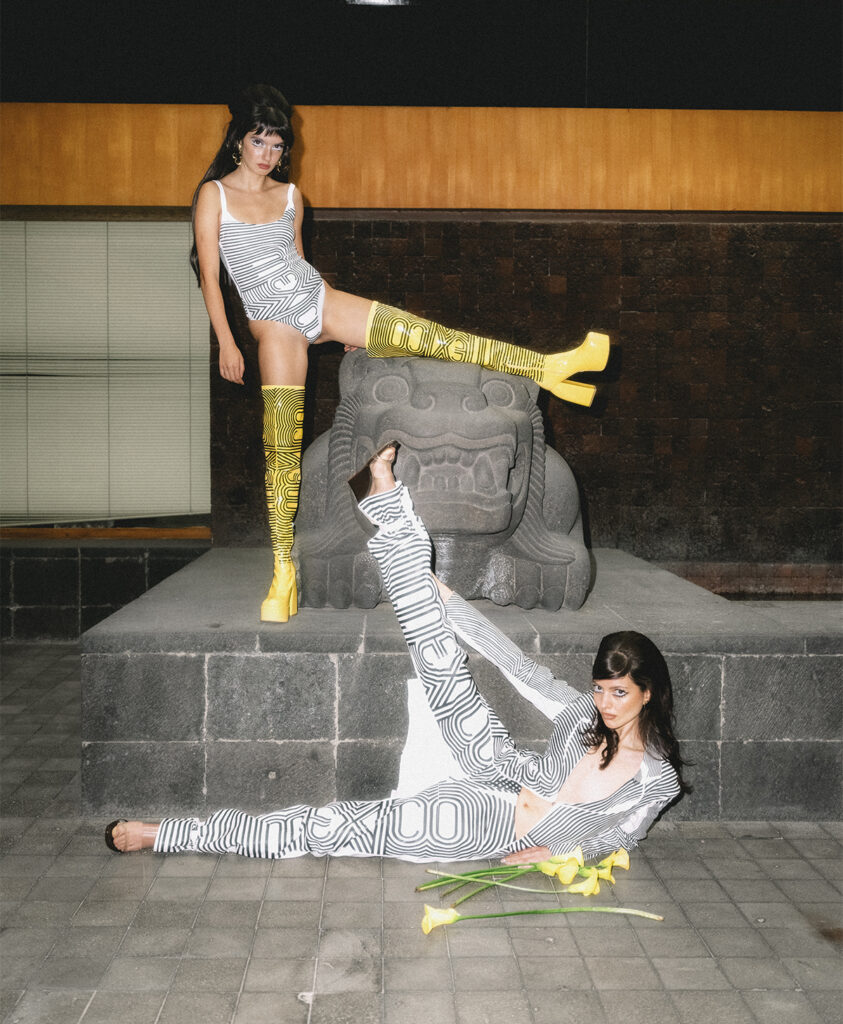 Dos modelos lucen prendas de la colección de Gabrielle Venguer inspirada en el lenguaje visual de México 68, posando junto a una escultura prehispánica en la casa de Pedro Ramírez Vázquez.