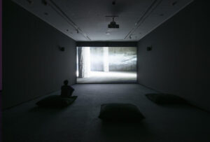 Sala de proyección con visitantes sentados sobre cojines frente a una videoinstalación que muestra un espacio arquitectónico envuelto en niebla y atravesado por un haz de luz.