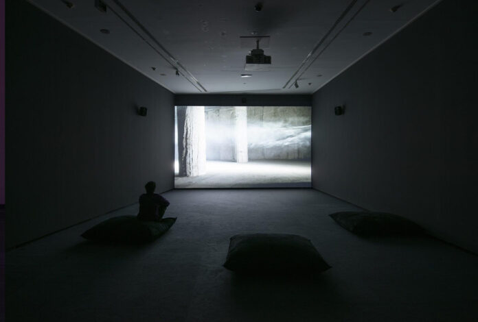 Sala de proyección con visitantes sentados sobre cojines frente a una videoinstalación que muestra un espacio arquitectónico envuelto en niebla y atravesado por un haz de luz.