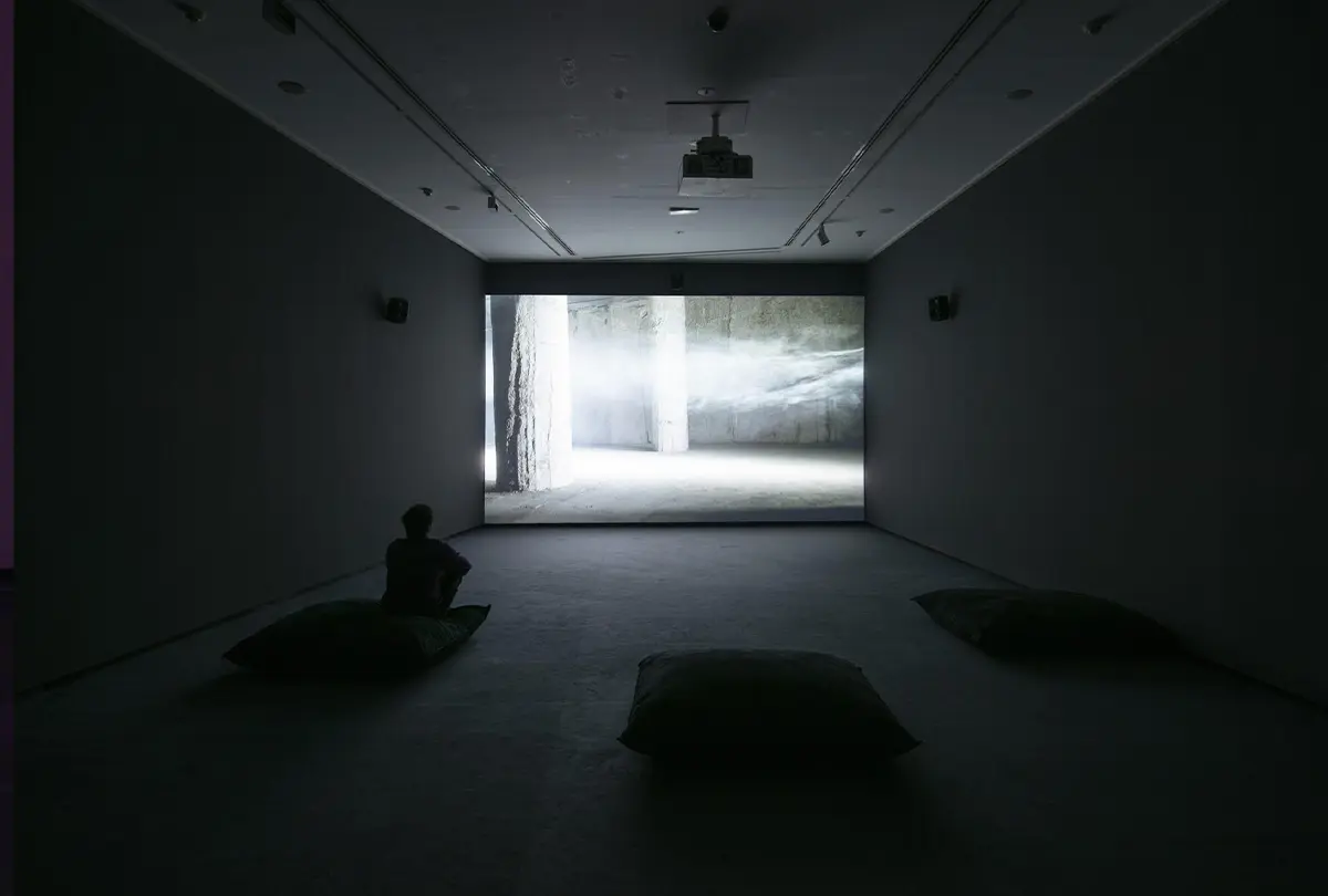 Sala de proyección con visitantes sentados sobre cojines frente a una videoinstalación que muestra un espacio arquitectónico envuelto en niebla y atravesado por un haz de luz.