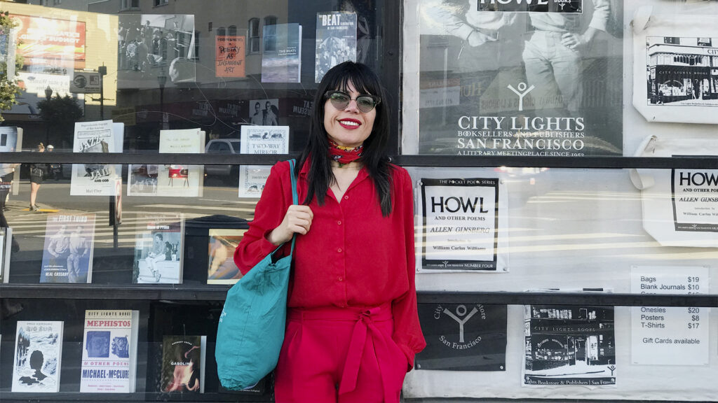 Sayak Valencia posa frente a la librería City Lights, en San Francisco, con ropa roja y una bolsa verde al hombro, sonriendo a la cámara.