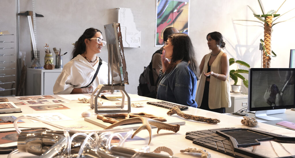 Un grupo de artistas y gestores culturales conversan alrededor de una gran mesa de trabajo con esculturas, maquetas y materiales experimentales en un showroom de Tijuana.