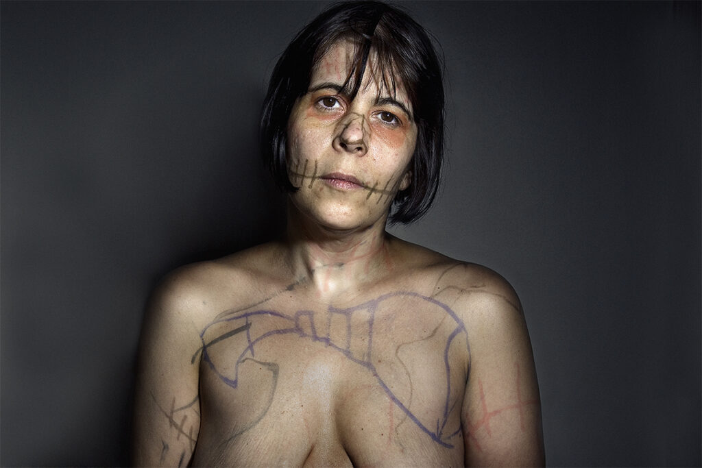 Retrato frontal de una mujer con el torso descubierto y dibujos y trazos sobre el rostro, el cuello, el pecho y los brazos, frente a un fondo oscuro.