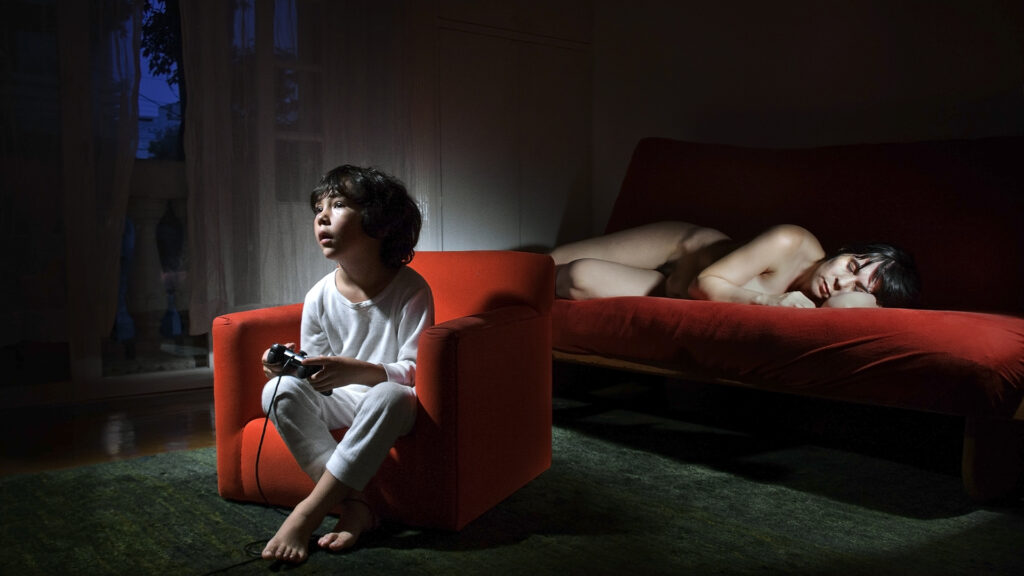 Niño sentado en un sillón rojo con un control en las manos, mientras una persona descansa en un sofá al fondo de una habitación en penumbra.