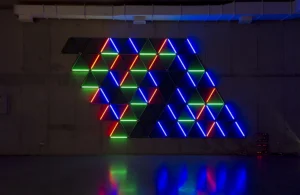 An installation by Gonzalo Reyes-Araos featuring triangular structures and tubes of red, green, and blue light against a dark wall.